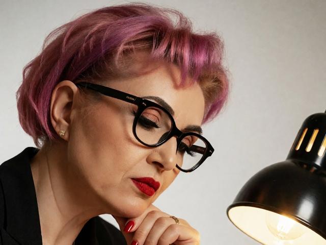 IsabelaConte with pink hair, black glasses, red lipstick, and red nails, looking down at a black lamp. Her hand is near her chin, and she's wearing a black shirt.