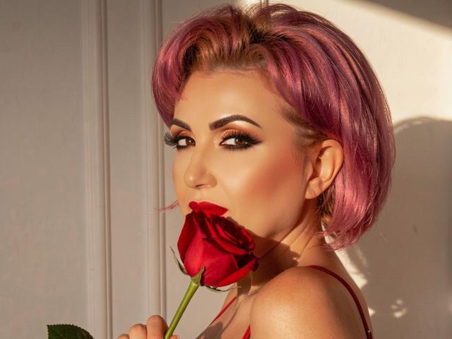 IsabelaConte with pink hair, holding a red rose to her mouth, has smoky eye makeup, red lipstick, and is looking seductively at the camera. Boobs partially visible.