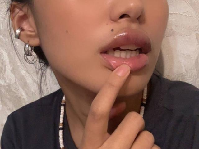 LeonilaSeewald's glossy pink lips are slightly parted with her finger touching the bottom one, showing a small mole above her upper lip. She wears a beaded necklace and hoop earrings.