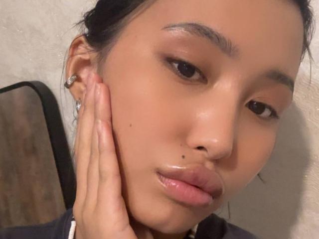 LeonilaSeewald's face with a small black mole on her left cheek, glossy pink lips, and her hand touching her cheek. She has brown eyes, black hair, and wears a silver earring.