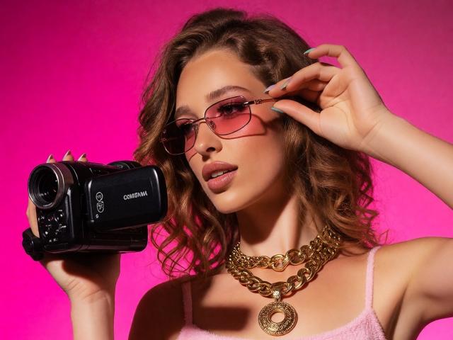 JessicaGibson holding a Canon camera, wearing pink glasses, a gold chain necklace, and a pink top, with wavy brown hair and teal nails, against a pink background.
