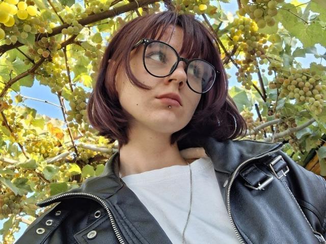 TeshaDotter mit kurzen, braunen Haaren, Brille, Nasenpiercing und Lippenpiercing, trägt eine schwarze Lederjacke über einem weißen Hemd und steht unter Weinreben..