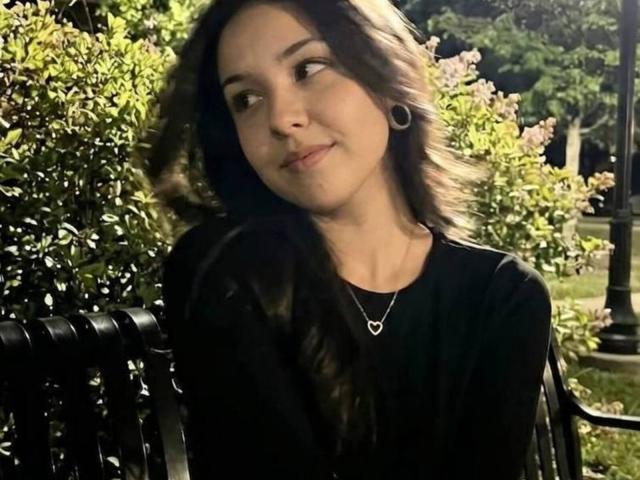 Debby Rubel, com cabelo castanho ondulado, pequenas argolas nos ouvidos, camisa preta e colar com um coração, sorrindo levemente, sentada em um banco de metal preto, com arbustos verdes e uma árvore ao fundo..