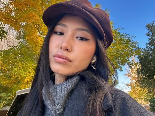 LeonilaSeewald with brown cap, long black hair, gray turtleneck, black coat, and earrings, pouting with subtle makeup, clear blue sky, autumn leaves, and sunlight in background.