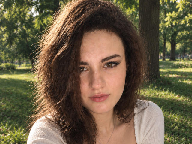 TreenaVisosky has curly brown hair, brown eyes, and a serious expression. She's wearing a white top, and her ass is not visible. The background is a sunlit park.
