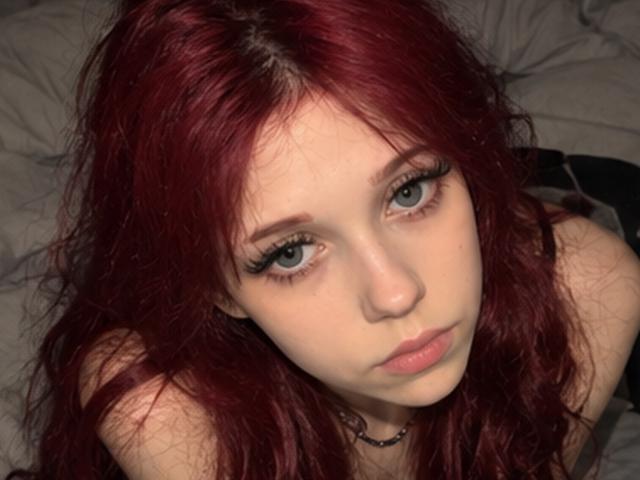 Elsa Caufield hat rotes, welliges Haar und blasse Haut, mit blauen Augen und langen Wimpern. Sie blickt nach oben, mit einem schmollenden Gesichtsausdruck, und trägt eine schwarze Halskette..