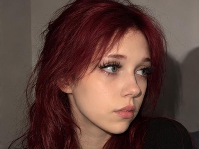Elsa Caufield, mit rotem Haar, heller Haut und blauen Augen, hat lange, schwarze Wimpern und einen leicht schmollenden Ausdruck, wobei sie leicht zur Seite schaut. Ihre Nase ist klein und niedlich..