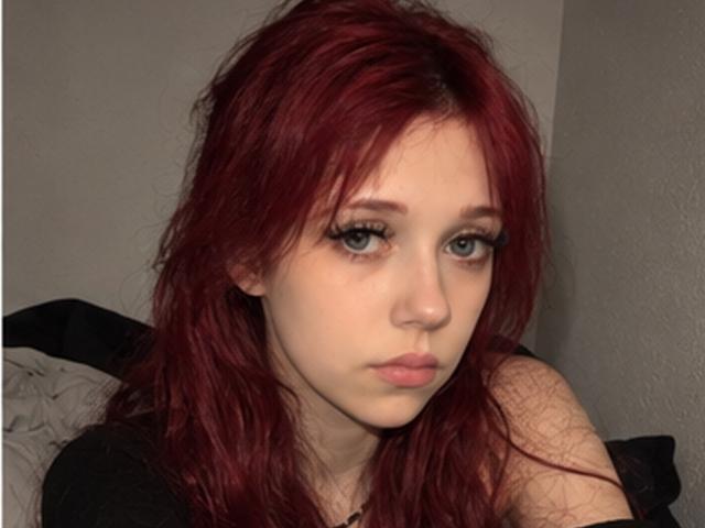 Elsa Caufield, con i capelli rossi, la pelle chiara e gli occhi verdi, ha labbra piene e lunghe ciglia, e sembra seria. Indossa una maglietta nera, con un braccio leggermente sollevato..