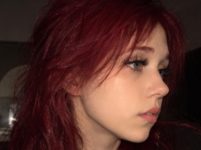 Elsa Caufield, con cabello rojo, ojos azules y largas pestañas, mirando hacia un lado con una expresión neutra, piel pálida y labios ligeramente rosados. Sus senos no son visibles..