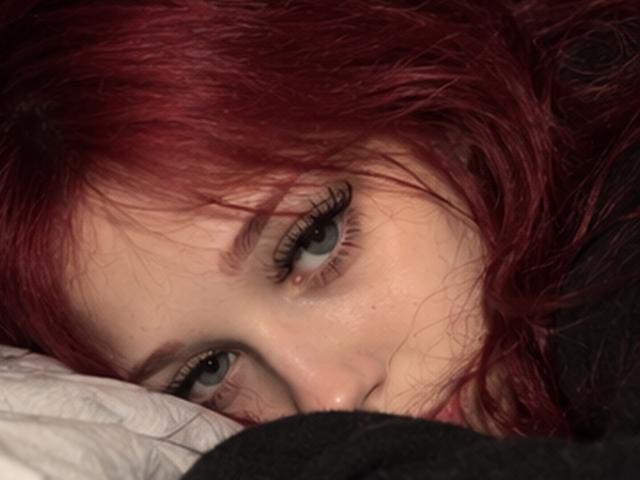 O rosto de Elsa Caufield, com cabelo ruivo e olhos azuis, olhando para cima com longos cílios escuros, deitada em um travesseiro branco, com uma roupa preta parcialmente visível..