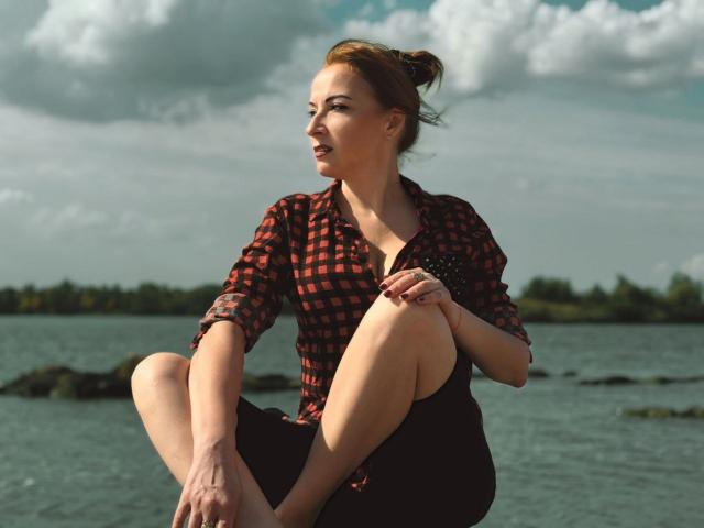ElizabethSo with red hair in a bun, wearing a red checkered shirt and black skirt, sitting with legs crossed, smiling, and looking to the side, by a lake.