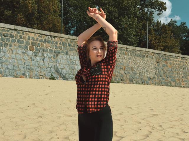 Elizabeth, les bras levés, portant une chemise à carreaux rouge et noire et un pantalon noir, debout sur un sol sablonneux, avec un mur de pierre et des arbres en arrière-plan..