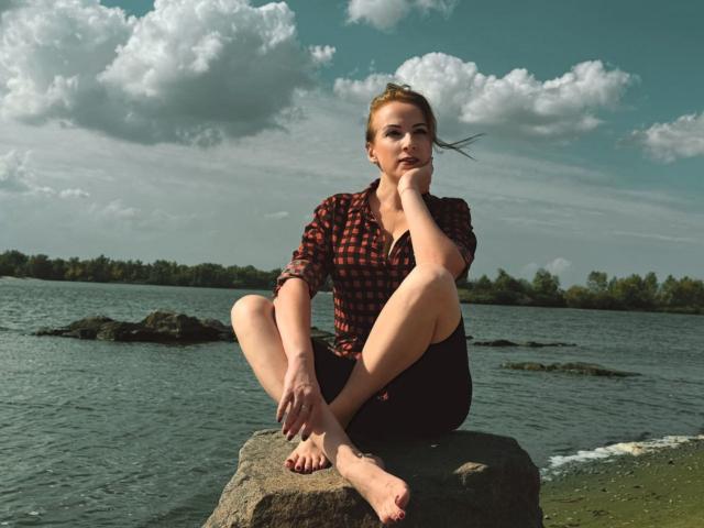 Elizabeth. Con los pies descalzos sobre una roca, vistiendo una camisa roja a cuadros y pantalones cortos negros, con un palo en la boca, y con una expresión pensativa. Sus pechos son pequeños..