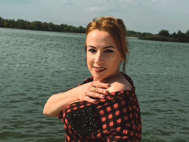 Elizabeth: Con i capelli rossi, indossa una camicia a quadri rossa e nera, con le mani sui seni, sorride alla fotocamera, in piedi in un lago, giornata di sole, indossa un anello..