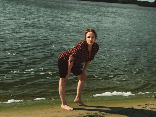 ElizabethSo, barefoot, bending forward with hands on knees, wearing a red plaid shirt and black shorts, standing on a sandy beach with greenish water in the background.