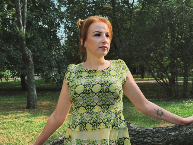 Elizabeth, con i capelli rossi, indossa un abito giallo-verde a motivi, è in piedi contro un albero, ha piccoli seni, un tatuaggio del sole sull'avambraccio destro e un'espressione leggermente infastidita..