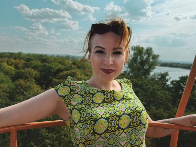 Elizabeth, mit dunklem Lippenstift, trägt ein grünes und gelbes Blumenkleid, hat die Arme auf einer Geländer, eine Sonnenbrille auf dem Kopf und ein selbstbewusstes Lächeln, draußen bei Tageslicht..