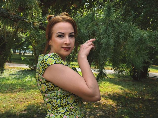 ElizabethSo with red hair, patterned green-yellow shirt, touching pine branch, smiling, outside, daytime, holding pine needle, green eyes, light skin, short hair, looking over shoulder, sexy.