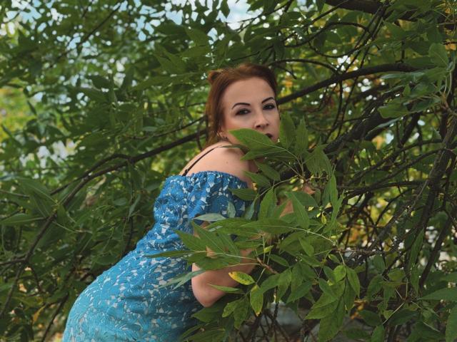 Elizabeth: Con i capelli rossi, il vestito di pizzo blu e i seni visibili, che spuntano tra le foglie verdi, mentre si guarda indietro con un sorriso seducente, circondata da una fitta vegetazione..
