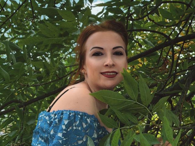 Elizabeth hat rote Haare, dunkle Augenbrauen und lächelt mit rotem Lippenstift. Sie trägt ein blaues Oberteil mit einem Schulterauschnitt, ist von grünen Blättern umgeben und hat einen schwarzen Träger..