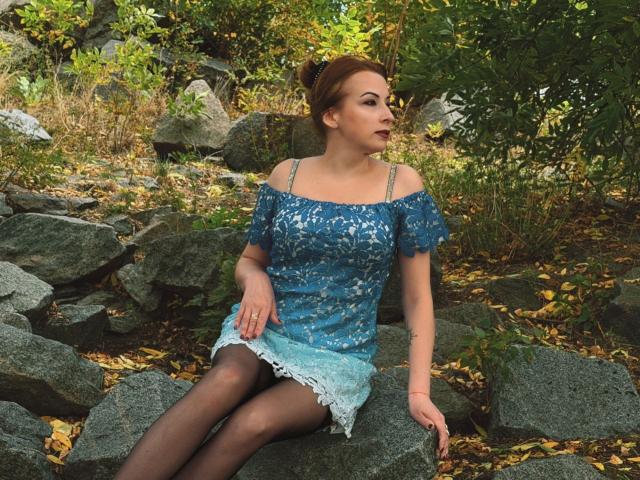 Elizabeth: Sentada em pedras, vestindo um vestido azul com um ombro descoberto e detalhes em renda, meias calças pretas e batom vermelho, com cabelo castanho preso em um coque, olhando para o lado..