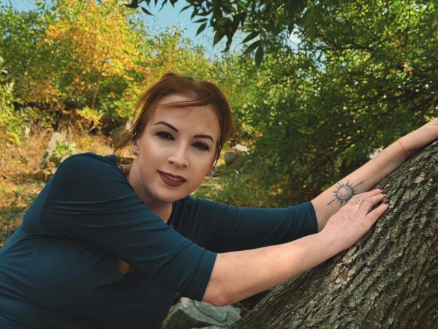 Elizabeth : Avec ses cheveux roux, sa chemise bleue et son tatouage de soleil sur le bras, elle est appuyée contre un arbre, souriante, montrant ses seins, dans une forêt. Elle a du rouge à lèvres foncé et un joli visage..