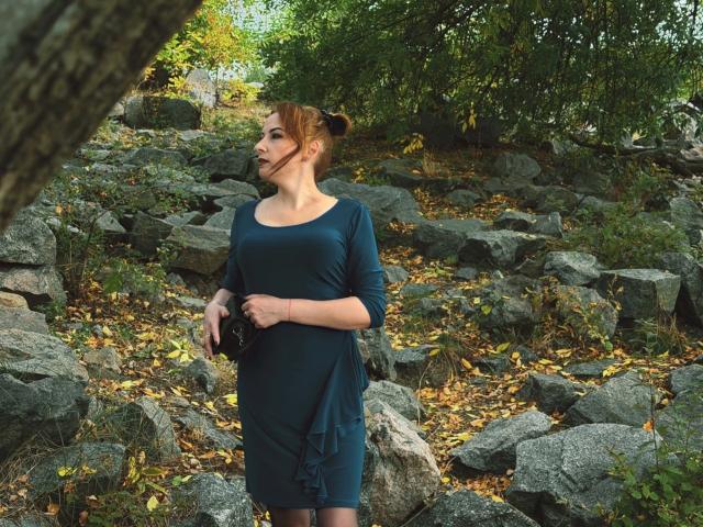 Elizabeth, con i capelli castani raccolti in uno chignon, indossa un abito color turchese, è in piedi tra le rocce, tiene una catena nera, guarda di lato, in una foresta con foglie verdi e gialle..