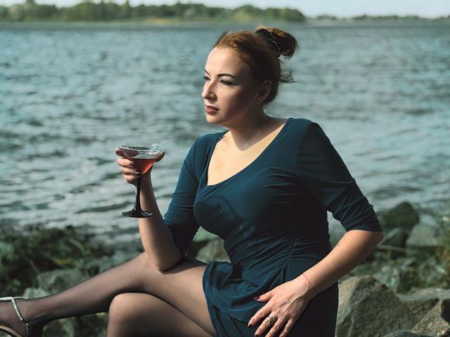 Elizabeth, con i capelli rossi raccolti in uno chignon, indossa un vestito blu e collant neri, è seduta su delle rocce, tiene in mano un bicchiere di vino rosso, appare pensierosa, con uno sfondo di acqua calma..