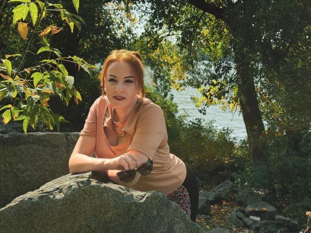 Elizabeth, met rood haar, leunt tegen een rots, gekleed in een roze shirt en luipaardprint broek, met een zonnebril in haar hand, lachend met donkere lippenstift, in een zonnig, bebost bos..