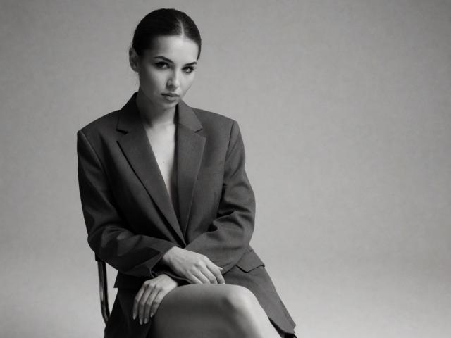 KayaOcean with dark, slicked-back hair, wearing a gray, buttoned-up suit jacket, sitting, legs crossed, no pants, exposing her bare, small, perky tits, serious expression, black-and-white photo.