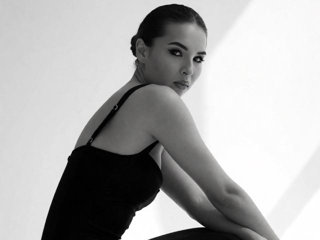 Black and white photo of KayaOcean with slicked-back hair, wearing a black tank top, sitting with arms crossed, looking over shoulder with a serious expression.