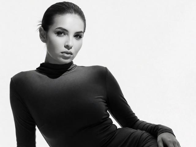KayaOcean with slicked-back dark hair, wearing a tight black turtleneck, sitting with a serious expression, her small tits subtly outlined by the fabric. Black-and-white, high-contrast.