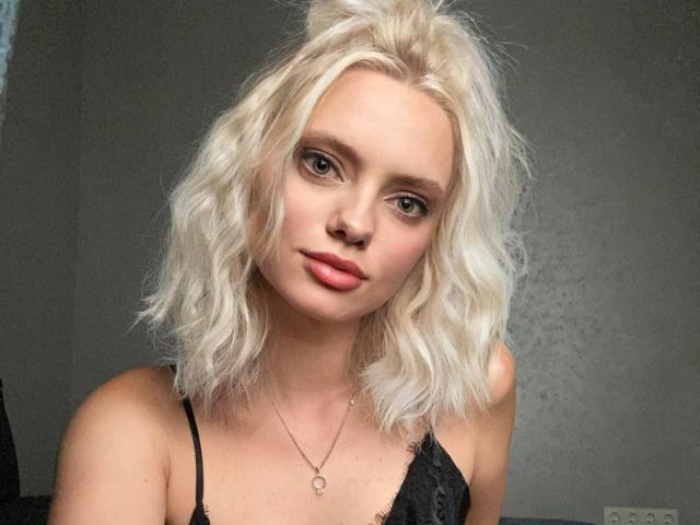 CrystalHope hat blondes, welliges Haar und grüne Augen und trägt eine schwarze Spitzenbluse. Sie schaut in die Kamera, hat rosafarbene Lippen, ein leichtes Lächeln und eine silberne Halskette..