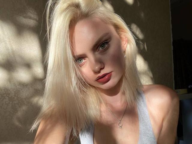 CrystalHope, com cabelo loiro, olhos verdes e batom vermelho, vestindo uma regata cinza, mostrando seus seios, em pé sob a luz do sol, olhando para a câmera com uma expressão séria..
