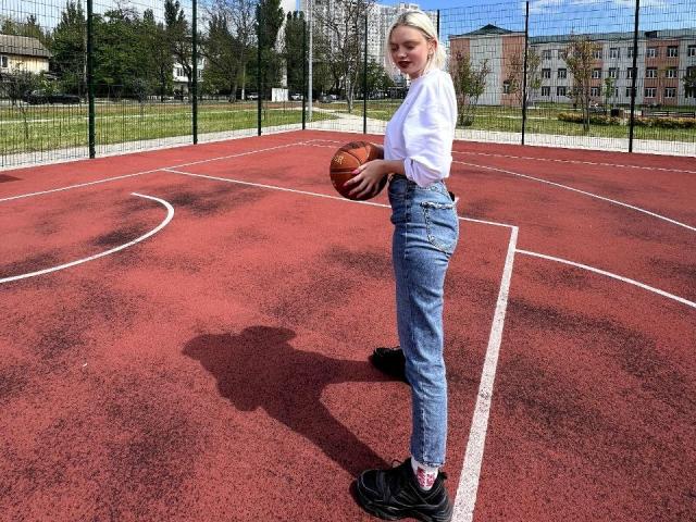 CrystalHope, con cabello rubio, vestida con una camisa blanca, jeans azules, zapatos negros, sosteniendo una pelota de baloncesto en una cancha roja al aire libre, con una sombra en el suelo, de pie de lado..
