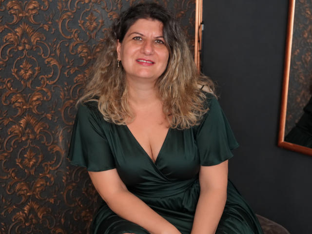 Roxy Vance, eine etwas mollige Frau mit lockigem, blondem Haar, trägt ein grünes Seidenkleid mit tiefem Ausschnitt und sitzt lächelnd. Ihre Brüste sind mittelgroß, und sie sieht niedlich aus..