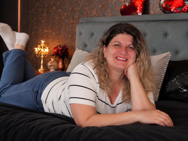 RoxyVance lies on a bed, legs up, wearing a white striped shirt and blue jeans, smiling with curly blonde hair, white socks, and a cute ass.