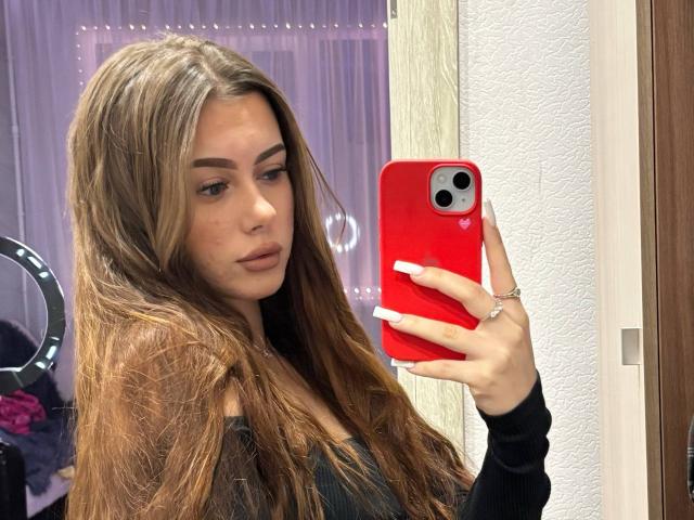 Lisabeth Lintner, met lang bruin haar, volle lippen en een rode telefoon, maakt een selfie, waarbij haar witte nagels en een ring te zien zijn. Ze draagt een zwarte top, en de foto is genomen in een spiegel..