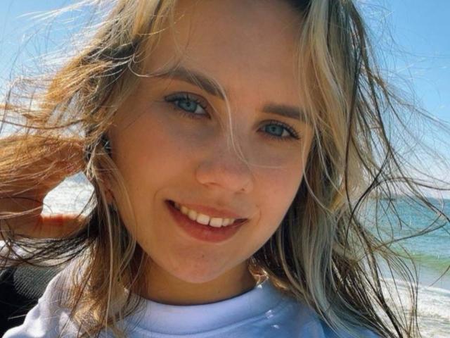 BettyLuv a les yeux bleus, les cheveux blonds et un sourire mignon avec les lèvres légèrement entrouvertes. Elle porte une chemise blanche, et ses cheveux sont emportés par le vent..