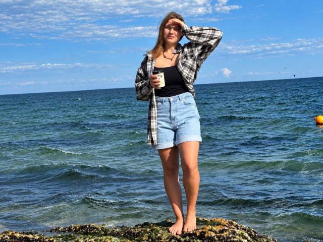 BettyLuv está descalza sobre la costa rocosa, vistiendo una camiseta negra, una camisa de cuadros y pantalones cortos vaqueros de cintura alta, sosteniendo una taza de café, con olas del océano y un cielo azul..