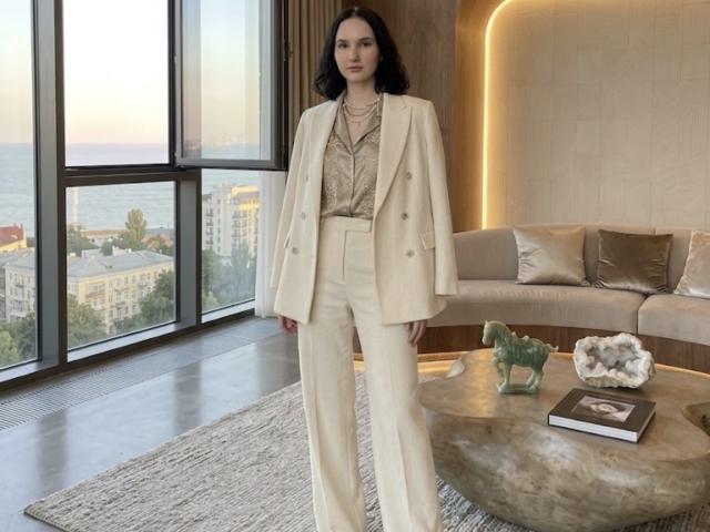 Ellaria has pale skin, dark wavy hair, and wears a cream double-breast jacket with high-waisted pants, a patterned blouse, and a necklace. She stands in a modern room.