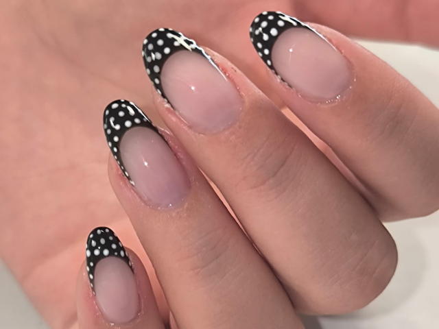 As unhas de ElvinnaLaFrancaise são um rosa nude brilhante, com pontas pretas e pontilhadas. Seus dedos estão ligeiramente afastados, exibindo a elegante e decorativa arte nas unhas..