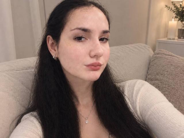 Ellaria with long black hair, pale skin, small nose, and pouty lips, wearing a white top and small earrings, sitting on a beige couch. Boobs not visible.