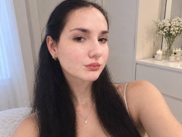 Ellaria with long black hair, light skin, small earrings, and a necklace, pouting her lips, wearing a white tank top, showing her bare shoulders and chest.