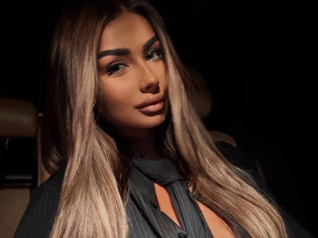LeylaBunny with long, straight, blonde-highlighted hair, dark eyebrows, full lips, wearing a black pinstriped shirt, looking slightly to the side with a subtle smile.