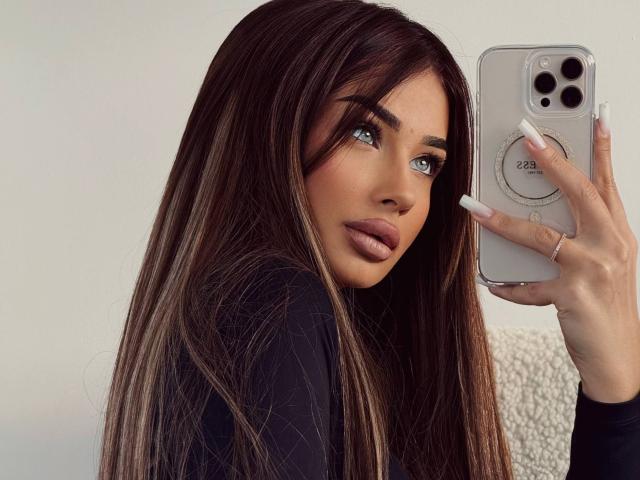 LeylaBunny with long brown hair, green eyes, and pouty lips, holding a phone, wearing a black top, white nails, and a ring, looking over her shoulder.