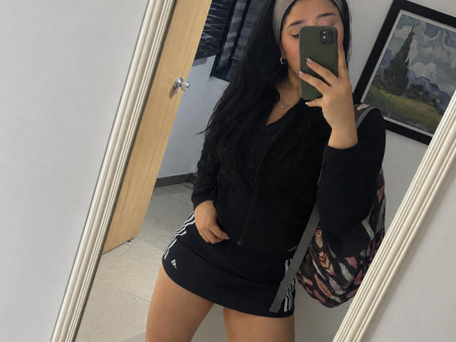 VickyMegan, com cabelos longos e pretos, vestindo um moletom preto e uma saia preta Adidas bem curta, tirando uma selfie no espelho, mostrando o bumbum e segurando um celular verde..