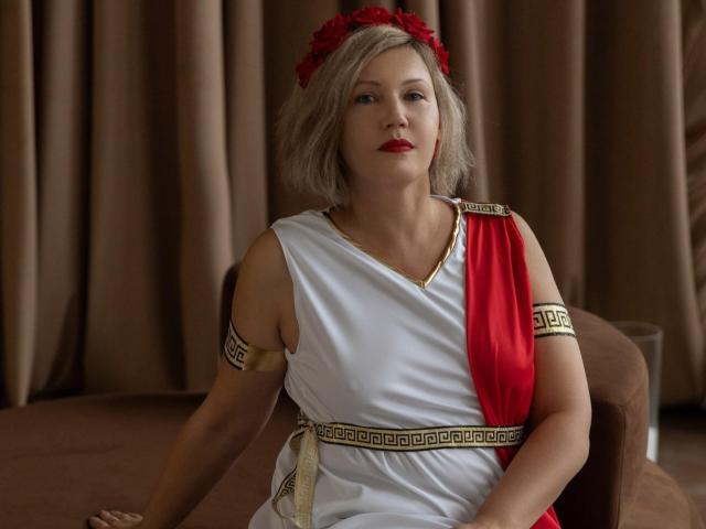 Nina Weiss, con cabello gris, una diadema de flores rojas, un cinturón rojo, un vestido blanco de estilo griego, brazaletes dorados y lápiz labial rojo, sentada en una silla marrón, con una expresión seria. Se pueden ver los senos debajo del vestido..