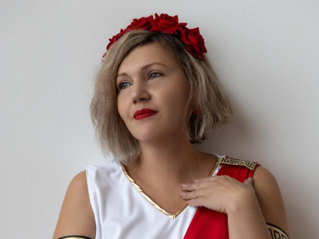 Nina Weiss mit blondem Bob, rotem Lippenstift und einem roten Blumenhaarschmuck, trägt ein rotes und weißes Kleid im griechischen Stil, eine Hand auf der Brust, blickt nach oben, weißer Hintergrund..