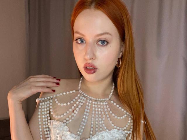 Janna Corsaro, avec ses cheveux roux, ses yeux bleus et ses lèvres rouges, portant un collier de perles sur son chemisier blanc en dentelle, sa main aux ongles rouges touchant son épaule..